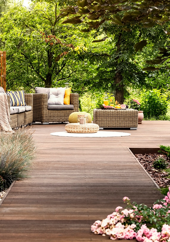 Terrasse en bois aménagée avec salon de jardin en osier, coussins colorés, table basse et poufs, entourée de verdure.