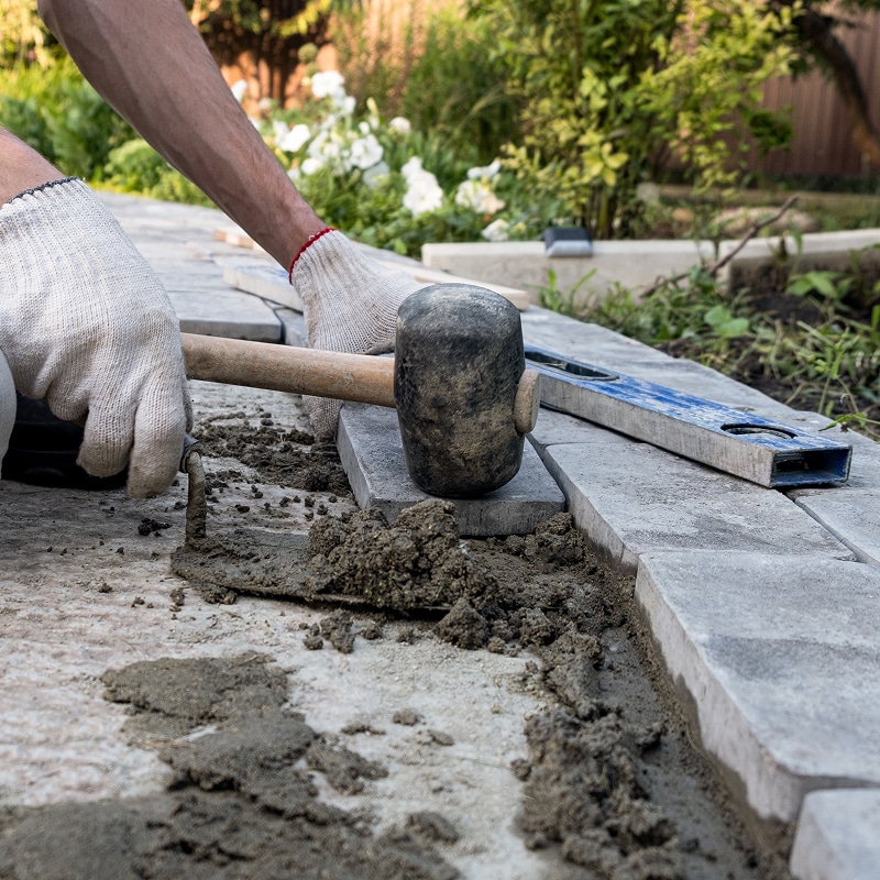 Pose de pavés dans un jardin. Mains gantées tapant un pavé avec un maillet en caoutchouc sur mortier. Nivelle visible.