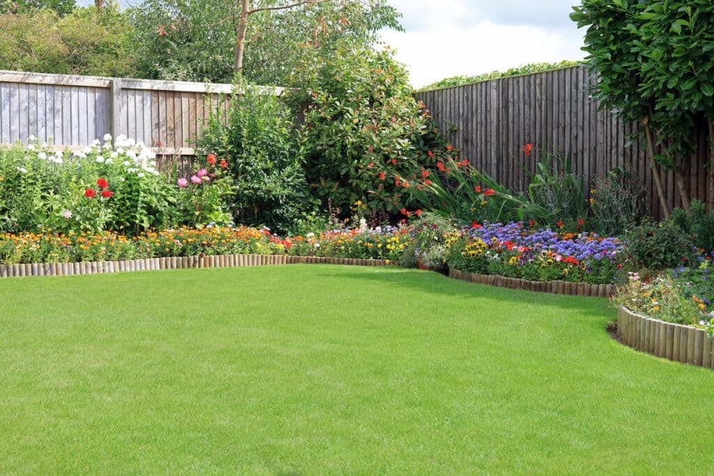 Jardin fleuri avec pelouse verte et clôture en bois. Massifs de fleurs colorées (blanc, rouge, orange, violet) le long de la bordure.